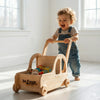 Smiling toddler pushes Balance mee wooden Montessori toy cart filled with soft toys in sunlit room.