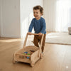 Smiling child pushing Balance mee wooden Montessori walker with blocks on hardwood floor.